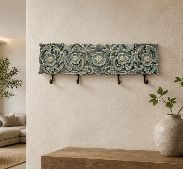 An ornate rectangular wall-mounted coat rack with carved floral details and five hooks, shown hanging on a beige wall in a styled interior with a vase and plants.
